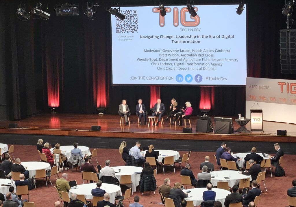 Stage with panelists at Tech In Gov 2024 Conference. Tables placed on conference hall floor in front of stage.