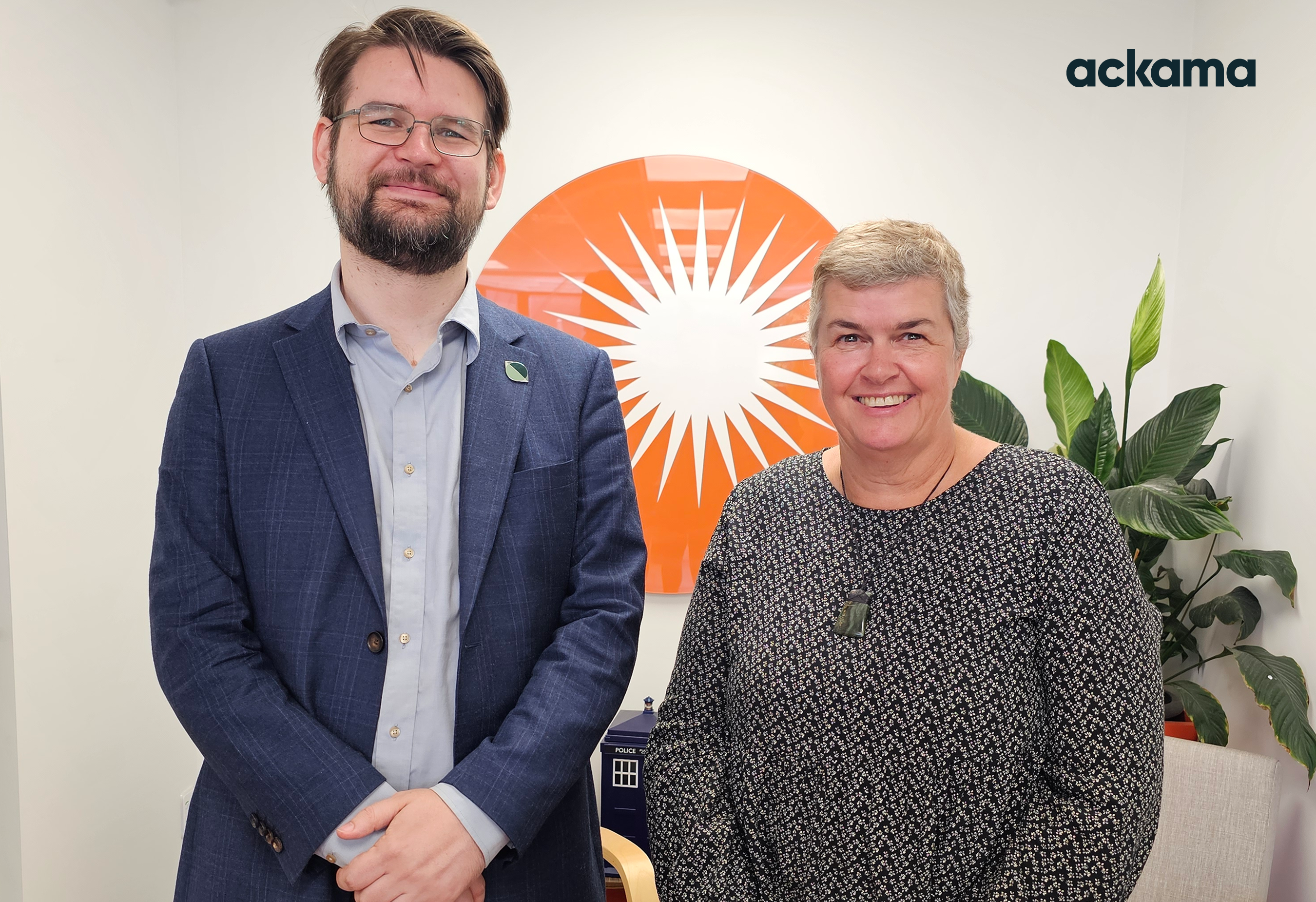 Two people standing side by side in an office setting, smiling at the camera. The man on the left has short brown hair, a beard, glasses, and is wearing a navy blazer over a light-blue shirt. The woman on the right has short, graying hair and is wearing a black patterned top with a green pendant. Behind them is an orange circular sign with white rays, and the word ‘ackama’ appears in the top right.